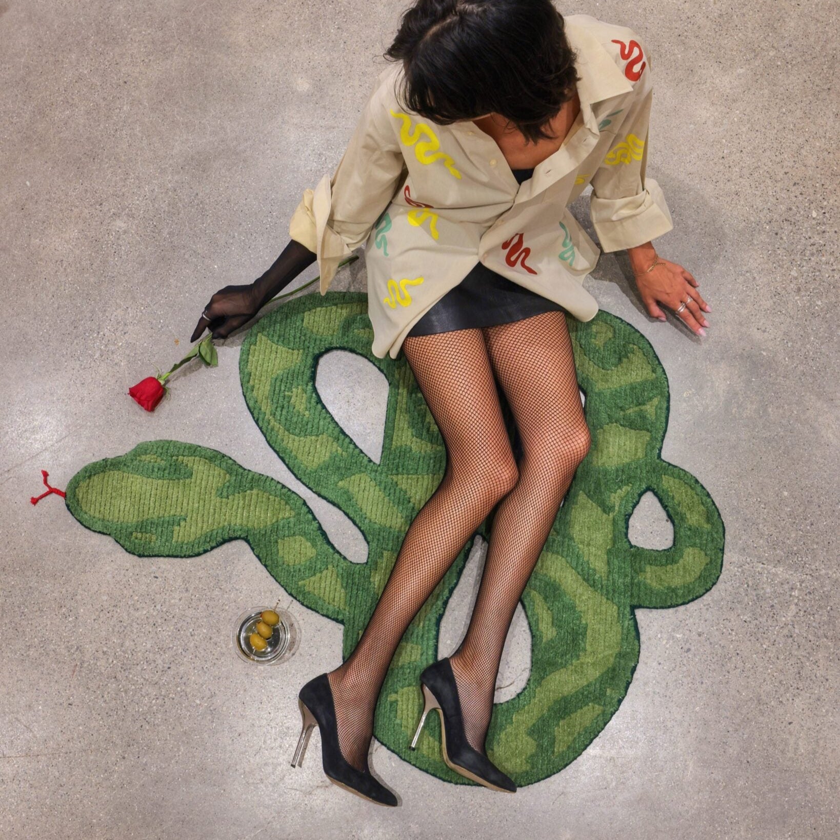 A contemporary artwork depicting a large green snake wrapped around a person lying on the floor, with the artist sitting beside the installation.