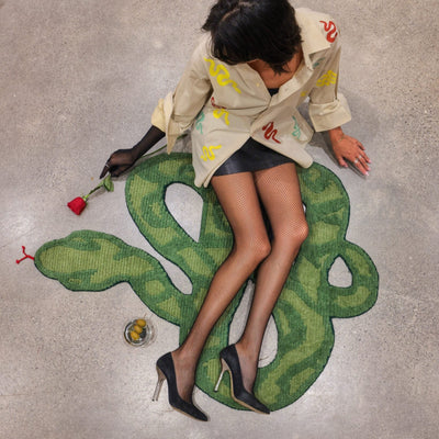 A contemporary artwork depicting a large green snake wrapped around a person lying on the floor, with the artist sitting beside the installation.
